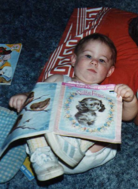 Dominick as a baby, lays on a pillow, with a book about a Puppy, open, in his hands. He is a baby with wide eyes, and is looking at the camera