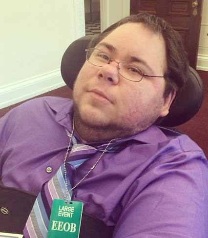 Dominick at the White House for a forum on LGBT and disability issues wearing a purple button down shirt, a purple and light blue striped tie, and a green Live Event badge, sitting in his black wheelchair.