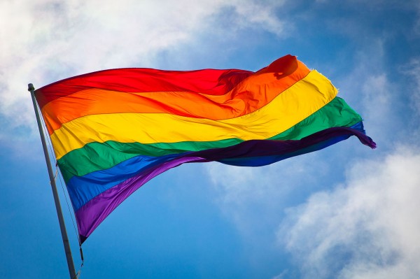 a rainbow flag flutters in the air, set in front of a blue sky.