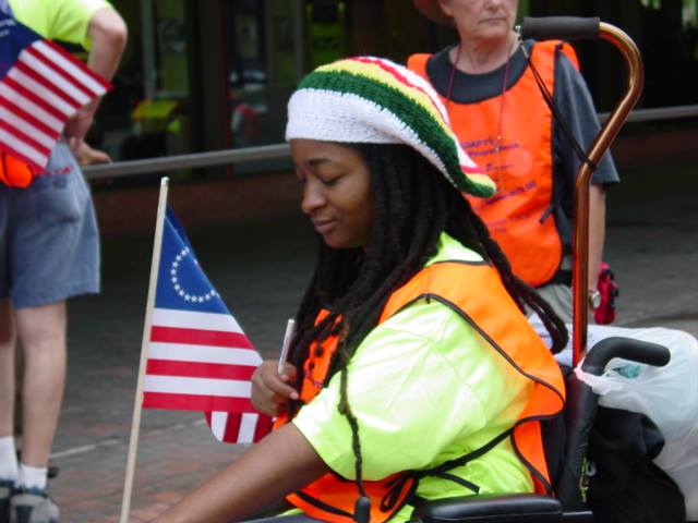 Anita Cameron in her Younger Days of protesting, over a decade ago she is wearing an orange vest, and sitting sideways with the disabled American flag behind her
