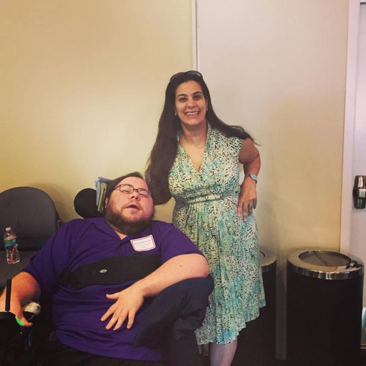 Maysoon is standing next to Dominick, her hand on his wheelchair, and other hand near her hip. She is wearing a green and white patterned dress. Her long dark hair is down around her shoulders, and she has a pair of dark sunglasses on her head. They are indoors. Dominick has a black power wheelchair. He is wearing a dark purple polo shirt, black pants, and has short dark hair and a dark brown beard.