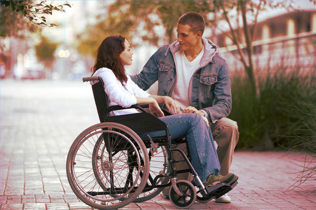  a woman sits in a manual wheelchair wearing a long-sleeved shirt and jeans with a man wearing a jacket kneeling down next to her and taking her hand