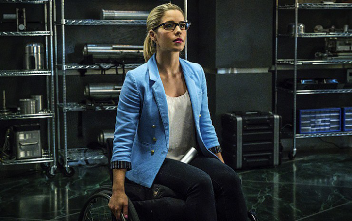 Emily Bett Rickards as Felicity Smoak sits in a manual wheelchair with her hands on the wheels. She is wearing a white shirt under an open blue shirt/jacket, and glasses and seems to be in a room with tech gear behind her