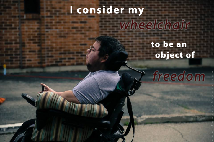 Dominick sits in his wheelchair on an outdoor film set, a red brick wall behind him. He is looking outward to the left, wearing a gray t-shirt, with a pillow propped under his arm. The image has the words I consider my wheelchair to be an object of freedom.