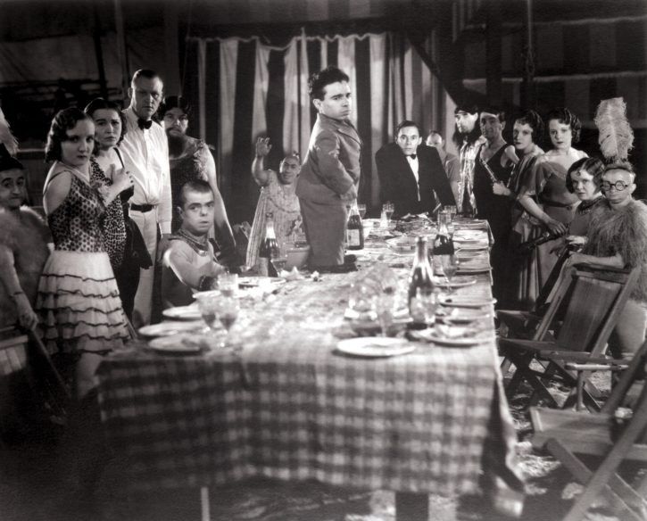  A scene from the movie Freaks by Tod Browning where a little person stands on a table covered in plates, food, glasses, etc. All of the disabled people are sitting around the table listening to him