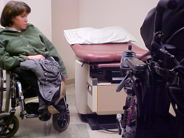 a woman in a wheelchair sits next to a small, empty examination table in a doctors office. It is a pink table with white paper crumpled on it. Another wheelchair user is sitting in the foreground on the right