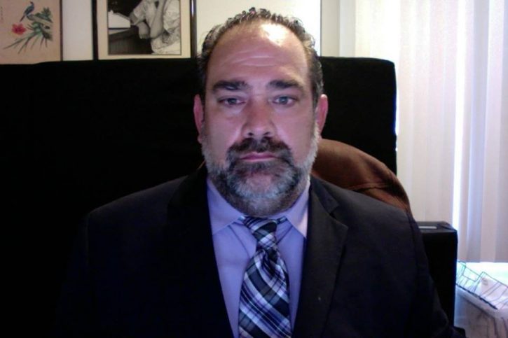  Daniel, a male identified individual,  sits in a leatherback  chair,  as he looks at the camera. He is wearing a suit and tie. His short hair is cropped back and he has a black beard with a hint of gray in it.