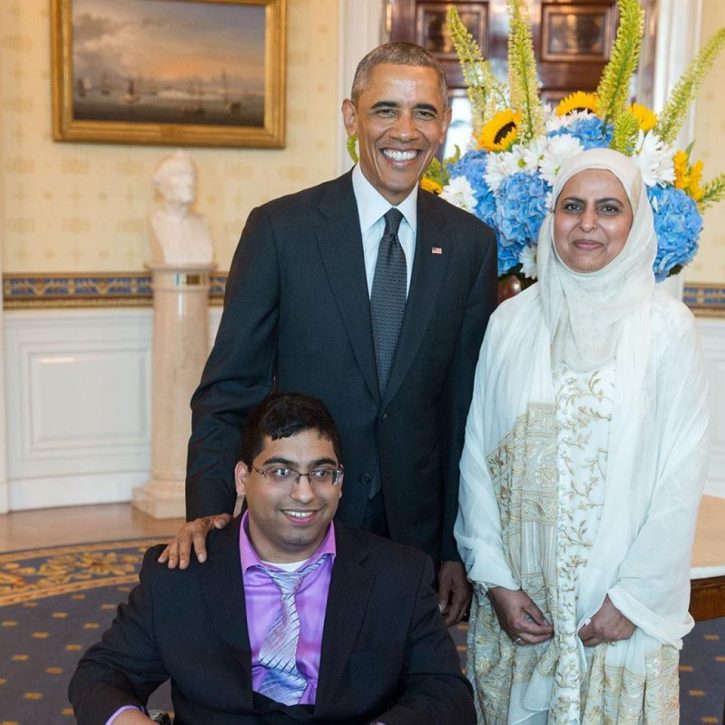 Hamza, a male identified individual sits in his wheelchair, wearing a suit, a smiling  Pres. Obama standing behind him, with his hand on Hamza's shoulder, and  he is next to Hamza's mother, a woman all in white, including a traditional looking Muslim hijab.