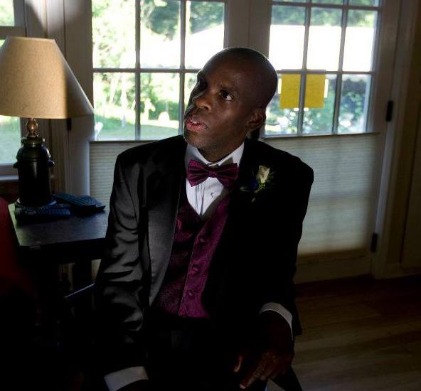 Leroy Moore, a black man with a disability, sits facing the camera. He is wearing a black suit with a bowtie, and a wall of windows is behind him