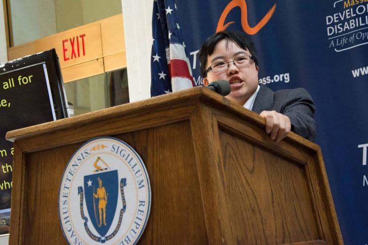 Young east asian person in business suit speaking at a podium. Podium has the seal of the Commonwealth of Massachusetts on it. There's a screen with CART captions beside the podium, and a banner with the logos for both the Massachusetts Developmental Disabilities Council and The Arc of Massachusetts.