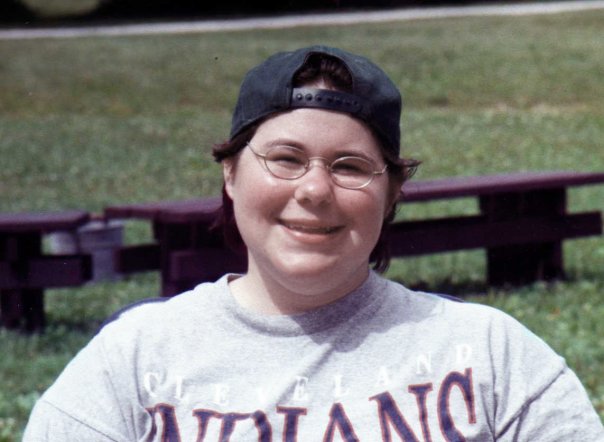 Dominick, as a teenager. A smiling face, wearing glasses, a background baseball cap, and a t-shirt, is visible from the shoulders up. There are picnic benches and grass in the background and only a little part of his wheelchair seat back is visible.