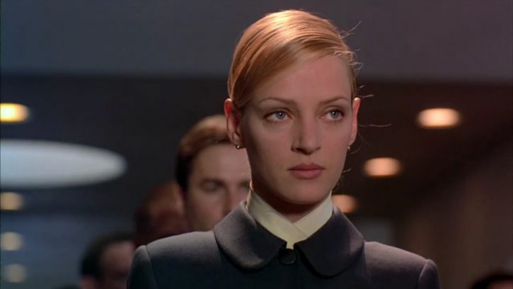 An image of Uma Thurman who plays Irene Cassini in the film, Gattaca. She is at debacle Corporation, wearing her uniform with her hair pulled back tightly. She has a serious expression on her face. People are standing behind her in a sort of line, and there are lights along the sides of the ceiling behind her.